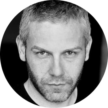 A black and white headshot of a man in his early fourties with a short head of hair, trimmed beard and black v-neck top staring at the camera