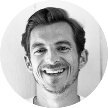 A black and white headshot of a man in his late thirties smiling at the camera with combed over hair and stubble