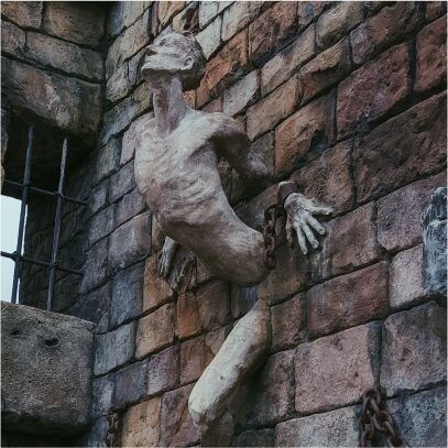 A side-view of a sculpture of an elderly man emerging from a wall, with shackles on his left wrist and chains along the bricks as he looks outwards, with a barred window in the corner brick wall gap in the foreground