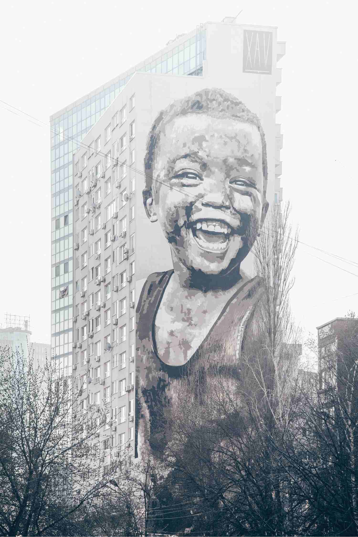 A large image of a young child, smiling at something in the distance, painted against a very tall block of residential flats on a foggy day with trees in the foreground