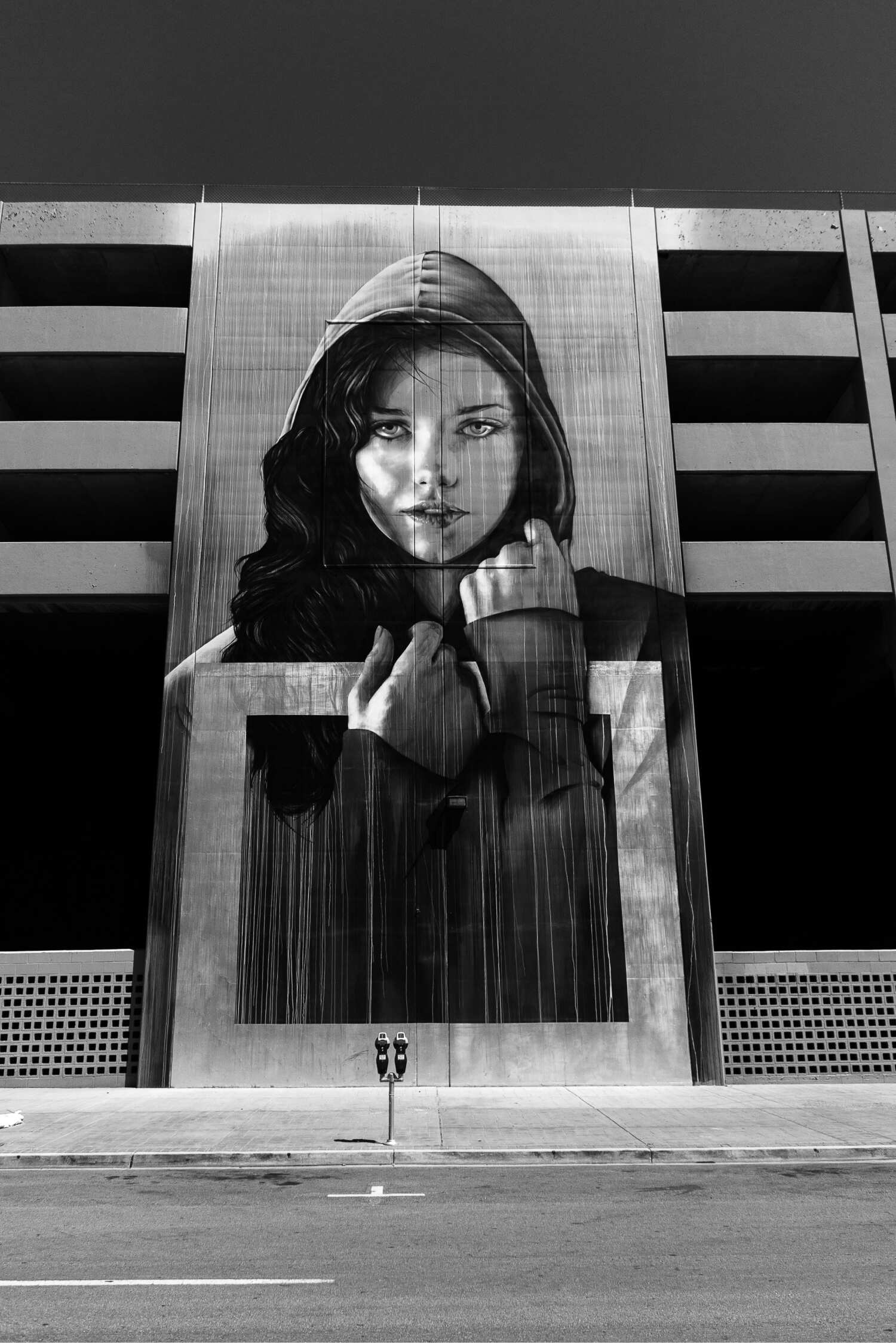 A mural of a young girl in a black hoodie on a tall face of a street building, looking at the viewer with her hands loosely clutching around the rim of her hood and neck, with long, dark, wavy hair flowing to the right (her perspective)