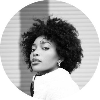 A black and white headshot of a young woman in her mid-twenties with decorative earrings and a white fleece staring at the camera, positioned sideways